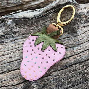 Puede incluir: Un llavero de cuero rosa en forma de fresa con un anillo de metal dorado y hojas de cuero verde. La fresa est&aacute; decorada con peque&ntilde;as cuentas rosas e iridiscentes.