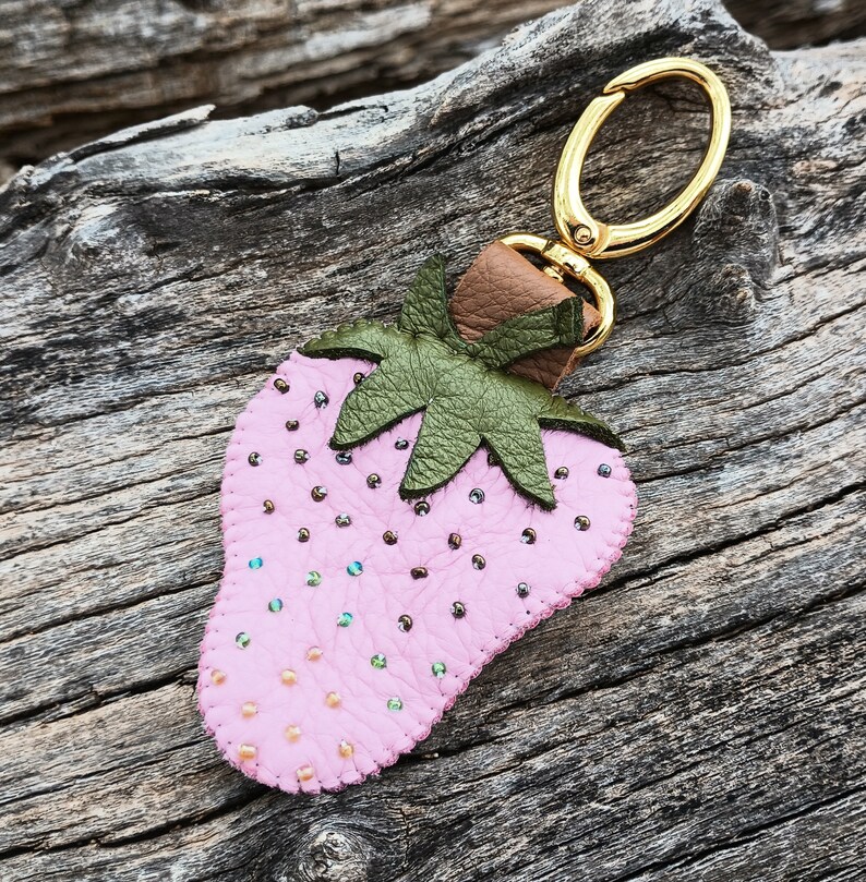 Puede incluir: Un llavero de cuero rosa en forma de fresa con un anillo de metal dorado y hojas de cuero verde. La fresa est&aacute; decorada con peque&ntilde;as cuentas rosas e iridiscentes.