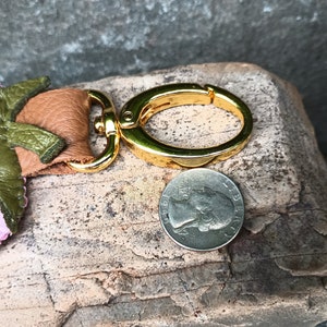 Puede incluir: Un llavero de fresa de cuero rosa y verde con un anillo de llave de metal dorado. El llavero es aproximadamente del tama&ntilde;o de una moneda de euro.