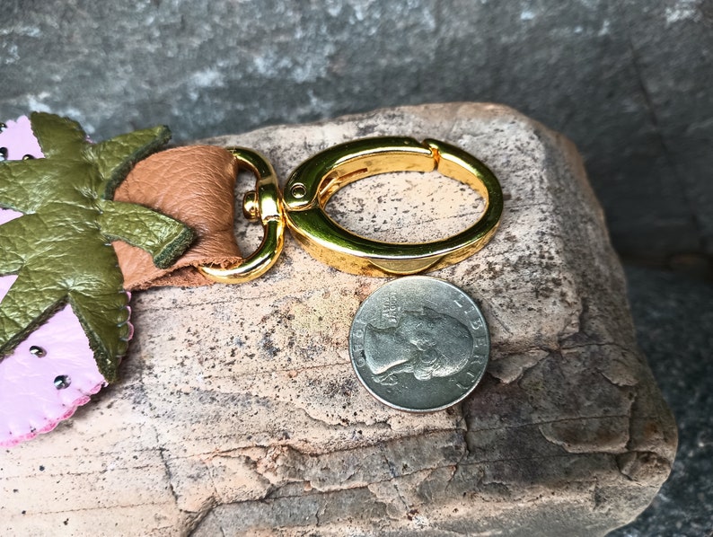 Puede incluir: Un llavero de fresa de cuero rosa y verde con un anillo de llave de metal dorado. El llavero es aproximadamente del tama&ntilde;o de una moneda de euro.