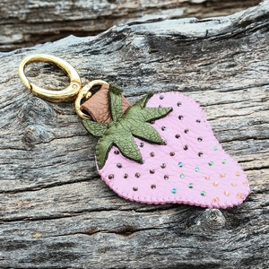 Puede incluir: Un llavero de cuero rosa en forma de fresa con hojas de cuero verde y un anillo de metal dorado.