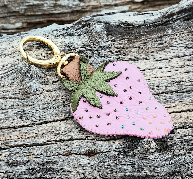 Puede incluir: Un llavero de cuero rosa en forma de fresa con hojas de cuero verde y un anillo de metal dorado.