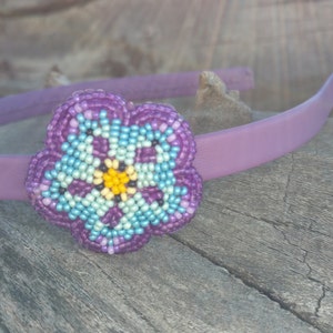 May include: A purple headband with a beaded flower accent. The flower is made of purple, blue, and yellow beads.