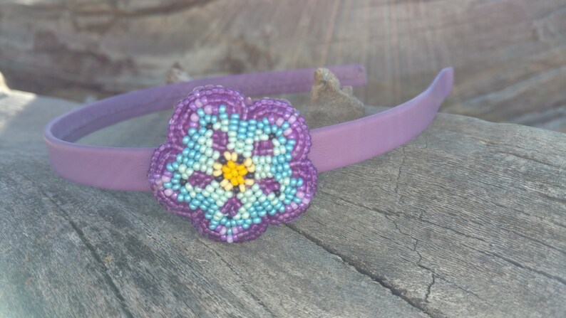 May include: A purple headband with a beaded flower accent. The flower is made of purple, blue, and yellow beads.