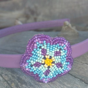 May include: Purple headband with a beaded flower accent. The flower is made of purple, blue, and yellow beads.