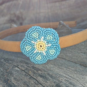 Puede incluir: Una diadema marr&oacute;n con un adorno de flor de cuentas azules y amarillas. La flor es un dise&ntilde;o de nomeolvides.