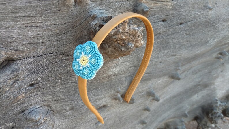 Puede incluir: Una diadema marr&oacute;n con un adorno de flor azul y amarilla. La flor est&aacute; hecha de cuentas y tiene un centro blanco.