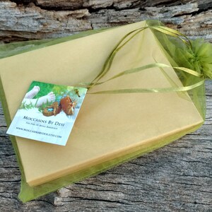 Puede incluir: Una caja de regalo verde y dorada con una cinta verde y una etiqueta que dice "Moccasins by Desi The Feel of Being Barefoot www.moccasinsbydesi.etsy.com"