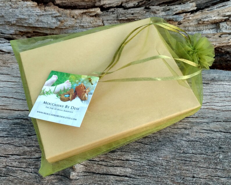 Puede incluir: Una caja de regalo verde y dorada con una cinta verde y una etiqueta que dice "Moccasins by Desi The Feel of Being Barefoot www.moccasinsbydesi.etsy.com"