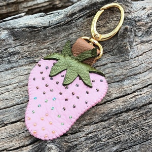 Puede incluir: Un llavero de fresa de cuero rosa con un anillo de metal dorado y hojas de cuero verde. La fresa est&aacute; decorada con peque&ntilde;as cuentas rosas e iridiscentes.