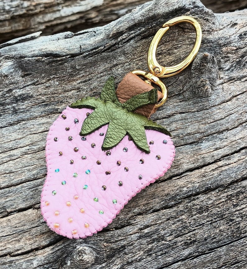 Puede incluir: Un llavero de fresa de cuero rosa con un anillo de metal dorado y hojas de cuero verde. La fresa est&aacute; decorada con peque&ntilde;as cuentas rosas e iridiscentes.