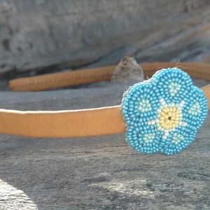 Puede incluir: Una diadema marr&oacute;n claro con una flor azul de cuentas. La flor tiene un centro amarillo y detalles blancos.