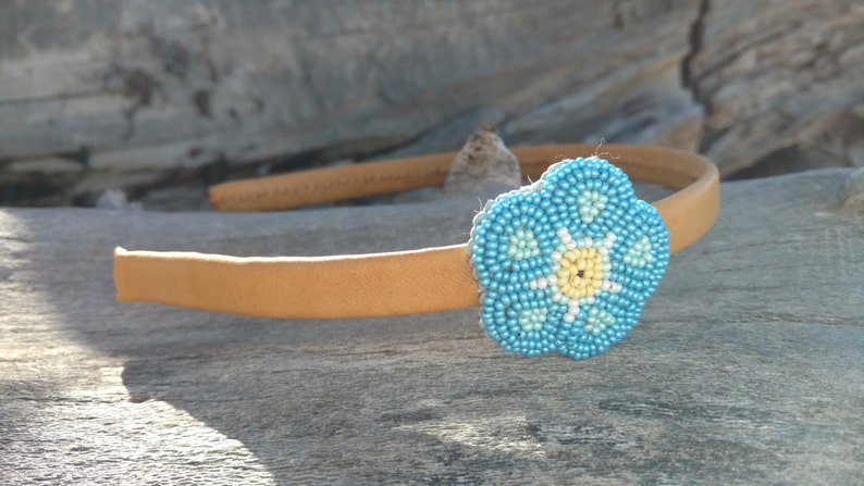 Puede incluir: Una diadema marr&oacute;n claro con una flor azul de cuentas. La flor tiene un centro amarillo y detalles blancos.