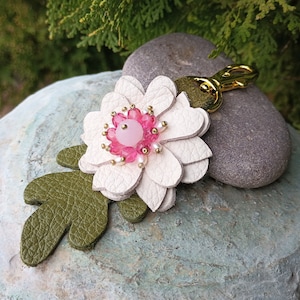 May include: A white leather flower keychain with pink and white beads in the center. The flower is attached to a green leather leaf and a gold metal key ring.