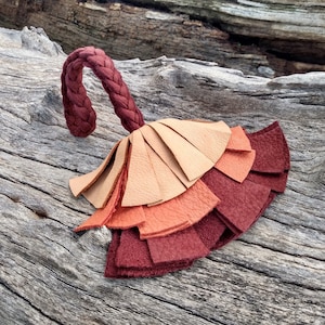 May include: A leather tassel with a braided leather loop. The tassel is made of multiple layers of leather in shades of brown, orange, and red.