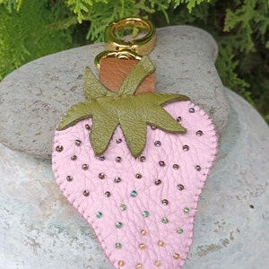 Puede incluir: Un llavero de fresa de cuero rosa con hojas de cuero verde y un anillo de metal dorado. La fresa est&aacute; decorada con peque&ntilde;as cuentas iridiscentes.