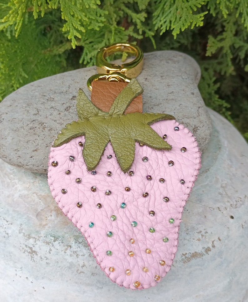 Puede incluir: Un llavero de fresa de cuero rosa con hojas de cuero verde y un anillo de metal dorado. La fresa est&aacute; decorada con peque&ntilde;as cuentas iridiscentes.