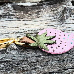 Puede incluir: Un llavero de cuero rosa en forma de fresa con una hoja de cuero verde y un anillo de metal dorado.