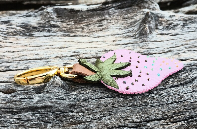 Puede incluir: Un llavero de cuero rosa en forma de fresa con una hoja de cuero verde y un anillo de metal dorado.