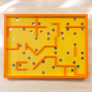 May include: A yellow and orange maze game with a green marble in the bottom right corner. The maze is made of orange plastic and has a yellow background. The marble is in the middle of a circle.
