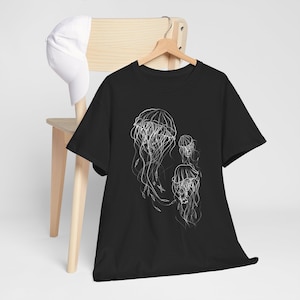 以下が含まれることがあります： 白い線画のクラゲが描かれた黒いTシャツ。デザインは、長い触手を持つさまざまなサイズの3匹のクラゲを示しています。シャツは木製のハンガーに吊るされています。