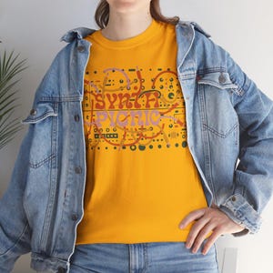 May include: Mustard yellow t-shirt with the text "SYNTH PICNIC" in a retro font. The design includes orange, pink, and green abstract shapes. The model is wearing a light blue denim jacket and jeans.