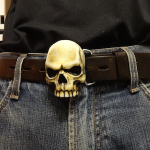 May include: A brown leather belt with a skull-shaped buckle. The skull is off-white with black eye sockets and a detailed jawline. The belt is worn with blue denim jeans.