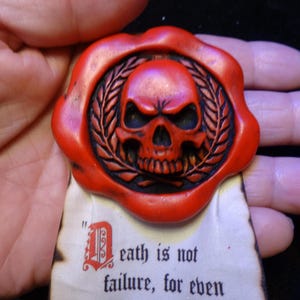 Puede incluir: Un sello de cera roja con una calavera rodeada por una corona de laurel, unido a un trozo de pergamino con bordes quemados. El texto en el pergamino dice: "Death is not failure, for even". El sello tiene un borde circular irregular.