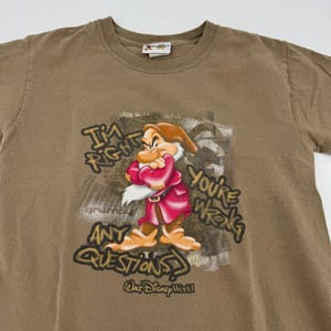 May include: A tan short-sleeved t-shirt featuring a graphic of the Disney character Grumpy with arms crossed. The text reads, "I'm right, you're wrong, any questions?" and "Walt Disney World" below the character on a distressed background.