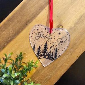 May include: Heart-shaped Christmas ornament with the words "Gledelig Jul" and a winter scene of trees and a cabin. The ornament is clear with black detailing and a red ribbon for hanging. The background is a wooden surface.