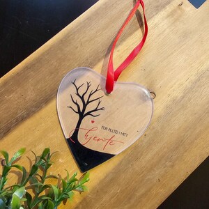 May include: Clear heart-shaped ornament with a black tree design and the word "Hjerte" in red script. A red ribbon is attached for hanging. The ornament is on a wooden surface.