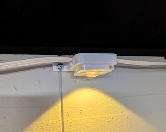 May include: A small white square LED light fixture mounted to a flat surface with a silver screw. The light is connected to a thin white electrical cord running horizontally and emits a warm yellow glow downward onto the surface below.