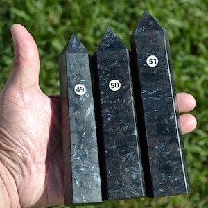 May include: Three black obsidian obelisks with white numbers 49, 50, and 51 on white stickers. The obelisks have a polished, reflective surface with subtle white and blue veining. The obelisks are held in a hand against a blurred green background.