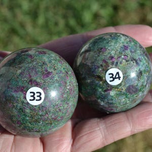 May include: Two green and purple gemstone spheres, numbered 33 and 34, are held in a hand. The spheres have a speckled appearance with a mix of green and purple hues. The background is blurred, suggesting an outdoor setting.