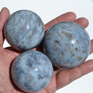May include: Three blue opal spheres with a marbled appearance are held in a hand. The spheres vary in size, with the largest one in the center. The spheres have a mix of blue and gray tones.