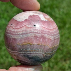 May include: A polished sphere of rhodochrosite, showcasing bands of pink, white, and gray hues. The gemstone ball is held by a person's fingers, with a green background. The sphere's surface displays a layered, wave-like pattern.