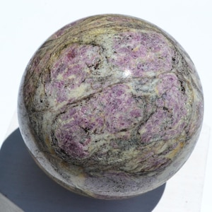 May include: A polished sphere with a marbled appearance. The stone features a mix of colors, including purple, gray, and cream. The sphere's surface has a smooth, reflective quality, and the overall effect is one of natural beauty.