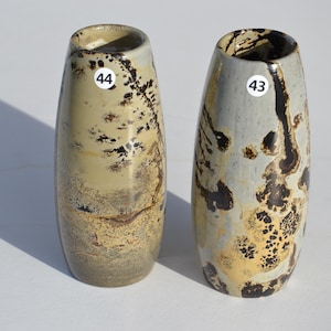 May include: Two cylindrical ceramic vases with rounded tops. The left vase, labeled 44, features a tan and brown mottled pattern. The right vase, labeled 43, displays a light gray surface with dark brown and gold-toned organic, spotted markings.