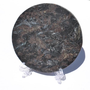 May include: A round, dark-colored stone with a textured surface and a mix of black, brown, and silver tones. The stone is displayed on a clear plastic stand, showcasing its natural patterns and mineral composition. The stone's circular shape is prominent.