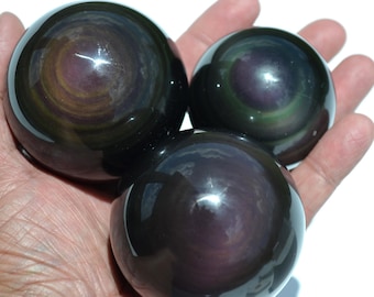 May include: Three polished obsidian spheres with iridescent swirls of color are held in a hand. The spheres have a dark, glossy appearance with visible patterns of purple, gold, and green. The overall theme is related to gemstones and crystals.