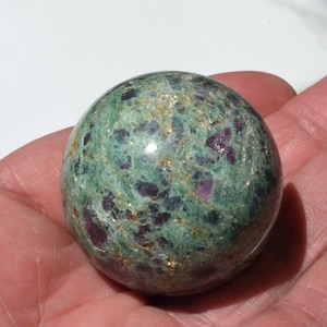 May include: A green, purple, and gold sphere made of polished ruby zoisite. The sphere is smooth and has a speckled appearance.