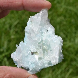 May include: A cluster of light blue and clear quartz crystals. The crystals have a rough, textured surface and are held in a person's hand. Small, dark specks are visible within the crystal formation. The background is blurred green.