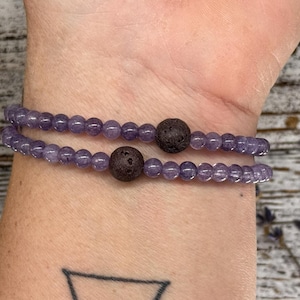 May include: A double-layered bracelet with purple beads and two brown lava rock beads. The bracelet is worn on a person's wrist.