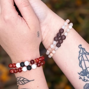 May include: Two bracelets with lava rock, white and red beads. The bracelets are stacked on each other on a person's wrist.