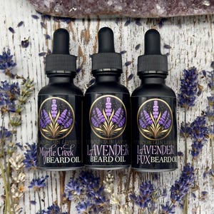 May include: Three small black bottles with gold labels and droppers. The labels read "Mountain Lavender Myrtle Creek Beard Oil", "Mountain Lavender Lavender Beard Oil", and "Mountain Lavender Lavender Fox Beard Oil". The bottles are arranged in a row on a white wooden surface with lavender flowers scattered around them.