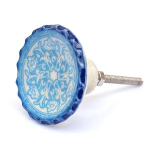 Op de afbeelding: Een ronde keramische meubelknop met een geschulpte rand en een blauw-wit bloemmotief. De knop heeft een glanzende afwerking en is voorzien van een metalen bout met een moer aan de achterzijde voor montage op meubels of kastdeuren.