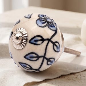 May include: A round ceramic cabinet knob with a cream-colored base featuring hand-painted blue floral and vine motifs. The center of the knob has a silver-toned metal cap with a fluted edge, and a threaded metal bolt extends from the back side.