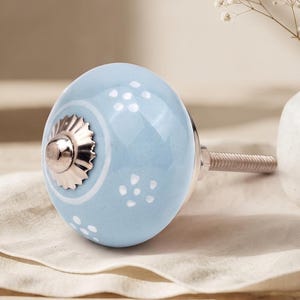 Puede incluir: Un pomo de cerámica redondo de color azul claro con un patrón floral blanco y una tapa central de metal plateado. El pomo incluye un perno de metal roscado que sobresale por la parte posterior para su instalación, con un acabado liso y brillante.