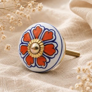 May include: A decorative ceramic cabinet knob with a floral design. The knob features a white base with a pattern of red and blue flower petals. A gold-colored metal center and screw complete the design. The knob is approximately 1.5 inches in diameter.