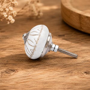 Op de afbeelding: Witte keramische kastknop met een zilveren basis en schroefdraad. De knop heeft een decoratief bladerpatroon in lichtbeige. De hardware is ontworpen voor meubels en interieurdecoratie.
